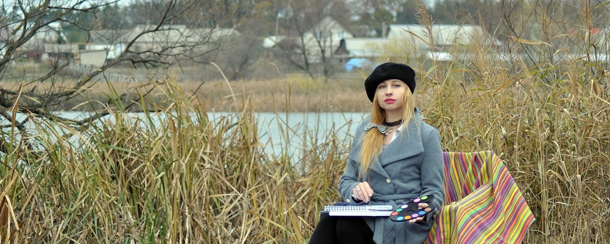 An attractive woman with long blonde hair sits on a folding chair on a river bank. She is wearing black boots, black leggings, a grey coat, and a black beret. She has a watercolour pad on her lap, a palette in one hand and brush in the other.