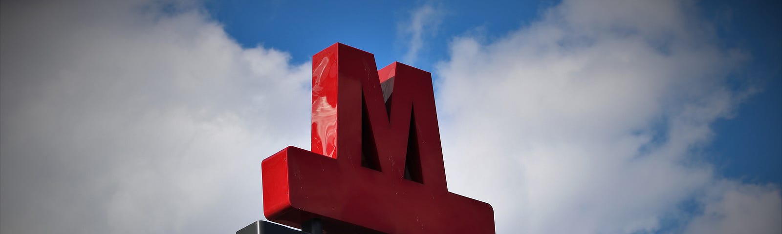 Large red M Copenhagen metro logo on a cloudy background with patches of blue