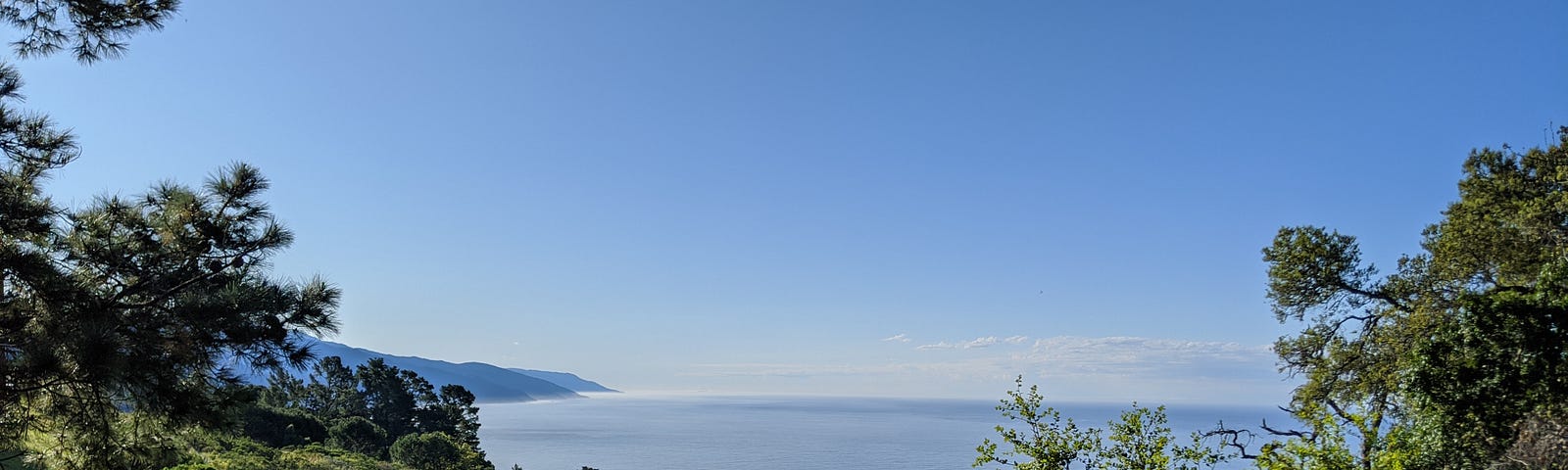 A photo of Big Sur, California, overlooking the Pacific Ocean and a mountain range in the background and a green hillside with trees in the foreground.