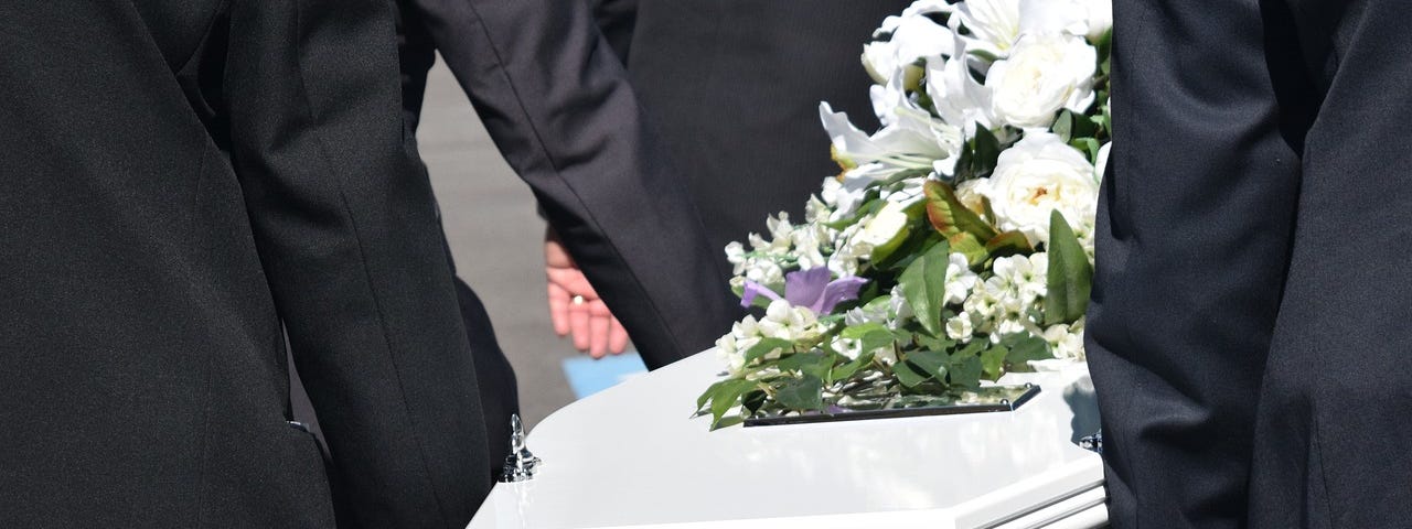 Image shows part of a white coffin in the center of the image, and parts of dark suits of people carrying it on the sides of the image.