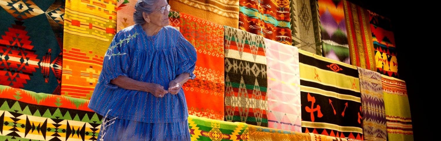 Collaged image of a Native American elder woman standing in front of colorful native blanket display