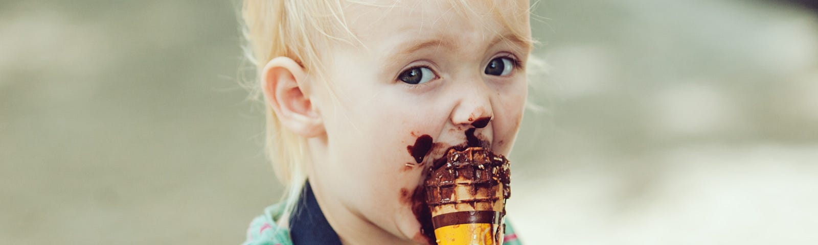 Toddler eating a chocolate ice-cream cone is looking right into the camera lens. He’s nose, cheeks, chin and hands are covered in chocolate ice-cream