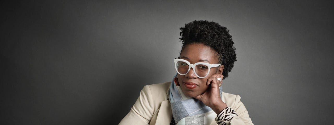 Joy Buolamwini smiles at the camera and wears white glasses. She is wearing a light beige blazer with a blue pattern scarf and a white shirt.