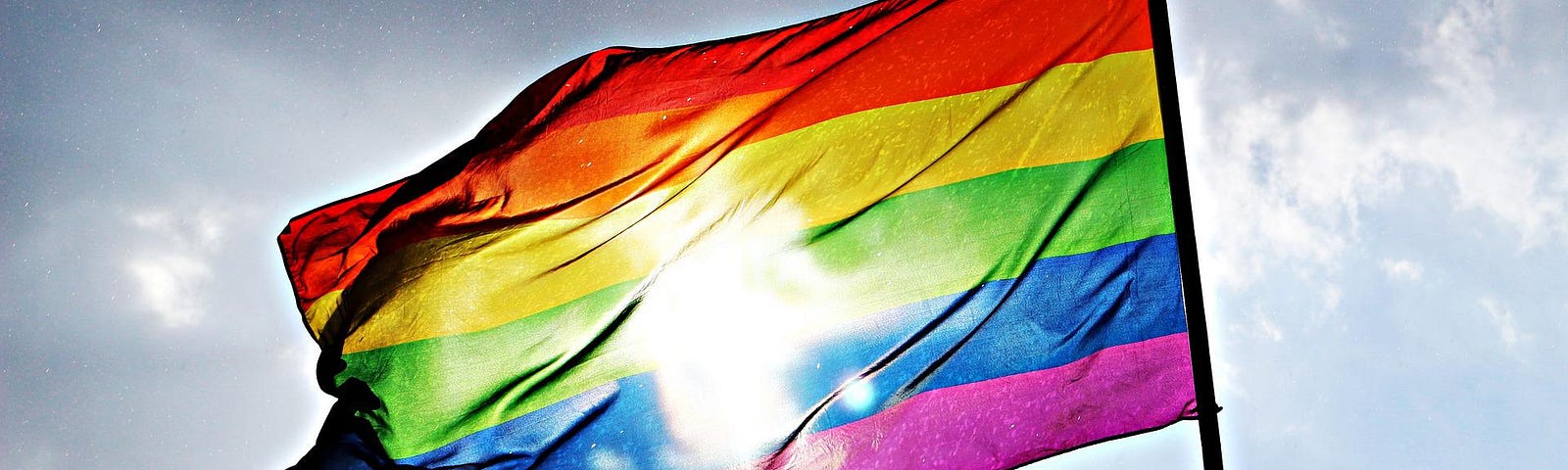 A rainbow pride flag flies against a blue sky with scattered white clouds. The sun is behind the flag, showing through the fabric.