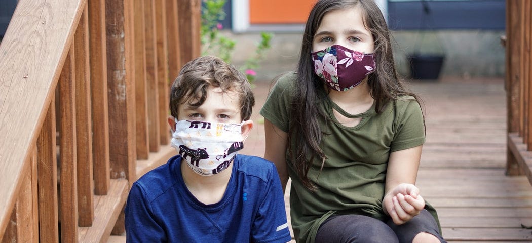Two kids wear masks while sitting on a porch.