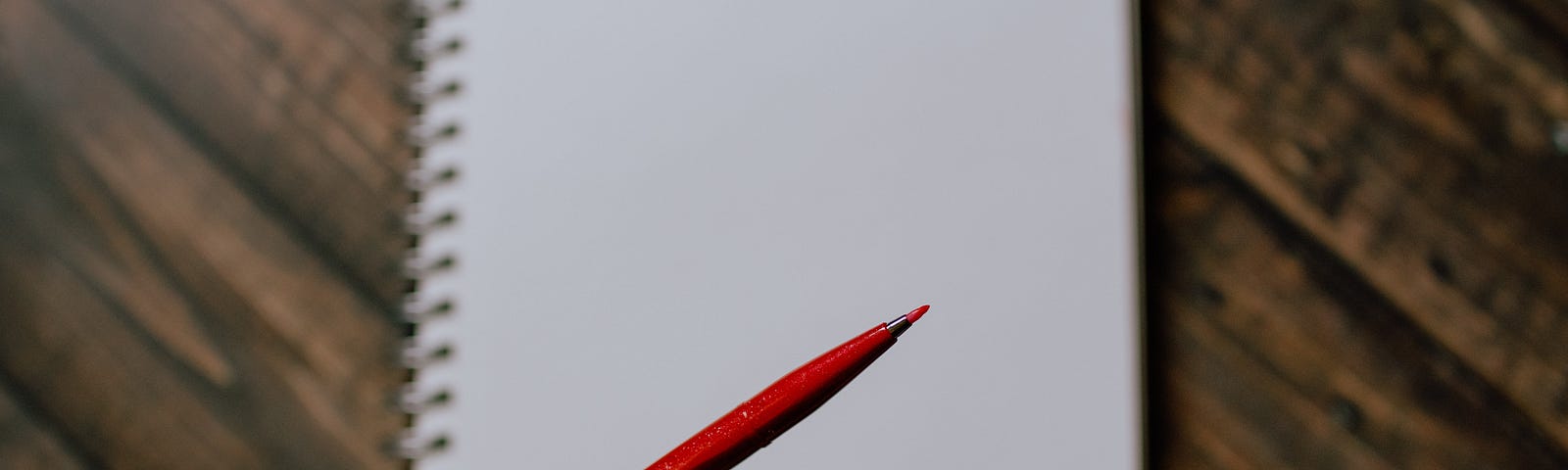 A left hand holds a red pen poised over a notebook on a wooden desk.
