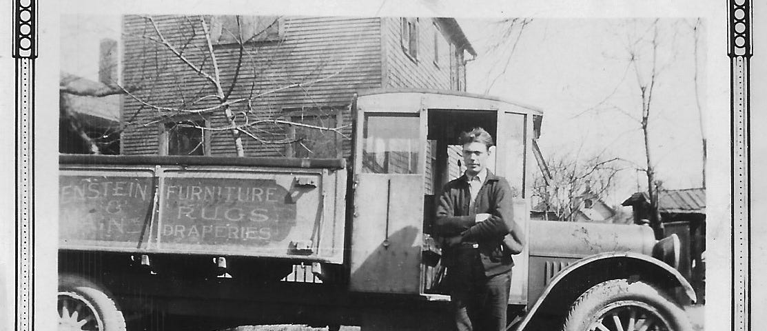 This shows a black and white photo of a man standing in front of a truck in the 1920s.