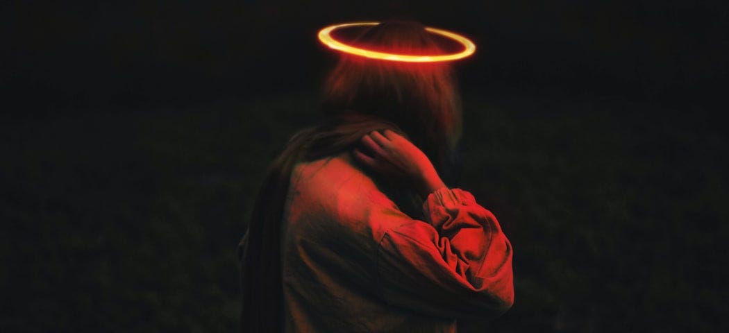 Photo of a person with their back turned. A glowing orange and red halo floats above their head, illuminating them in a deep red light. The background is black and blurry.