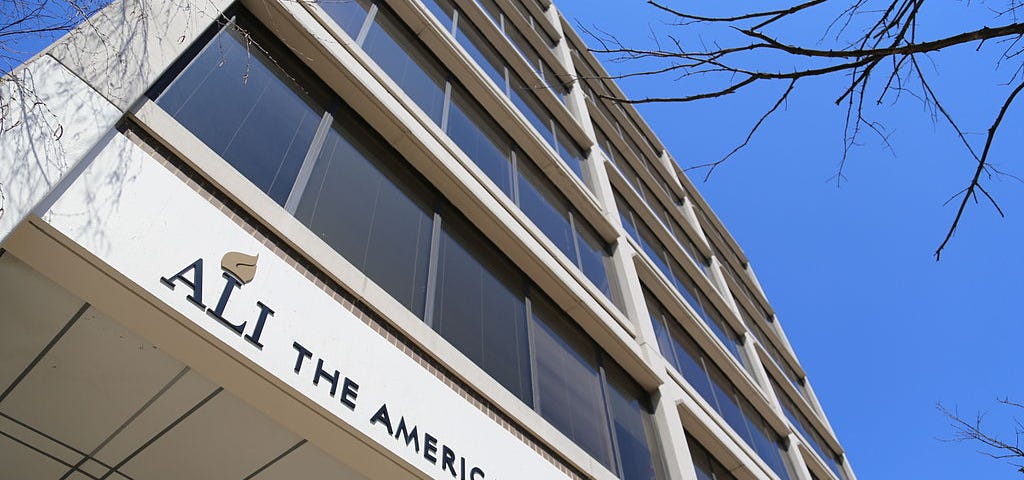 The front of an office building is shown in an upward perspective from the ground with a blue sky and some tree limbs in the background. The building has a sign over the entrance that reads “ALI THE AMERICAN LAW INSTITUTE.”