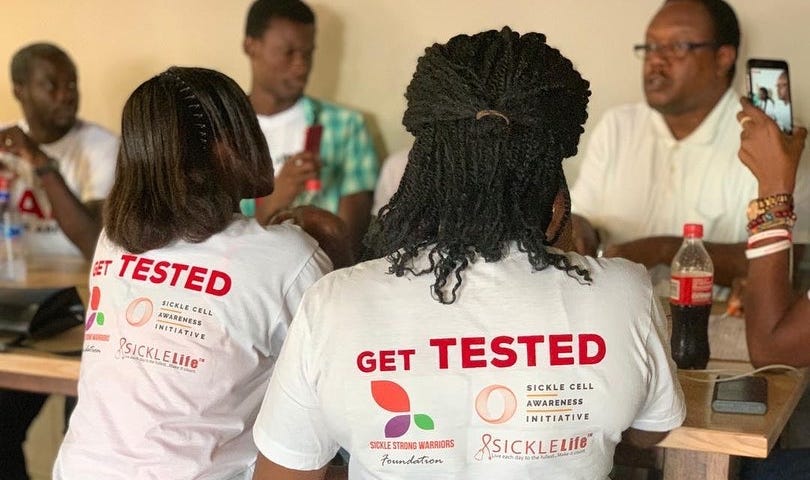 Two women wearing t-shirts reading ‘Get Tested’ sitting at a table face away from the camera, with other people out of focus in the background