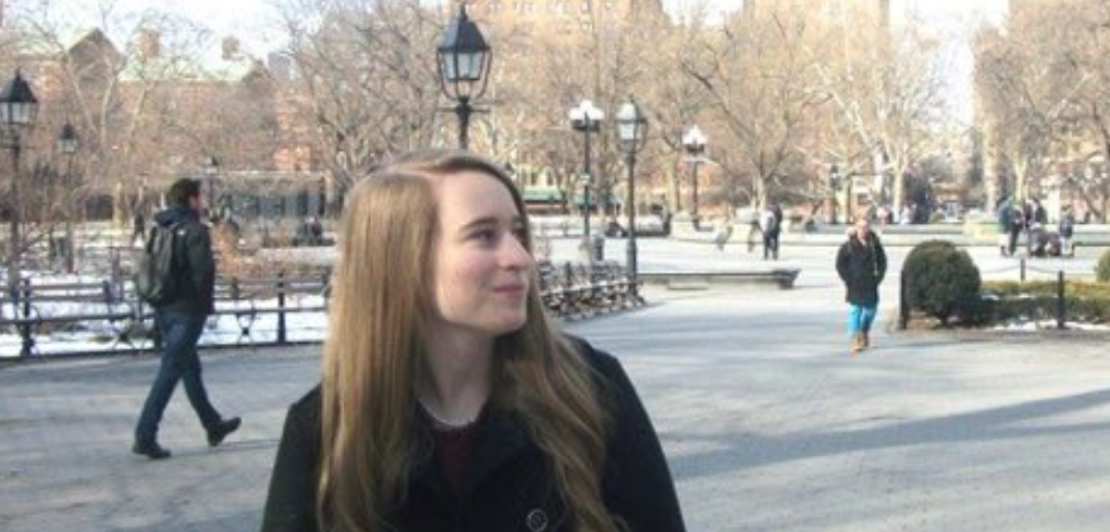 A girl with long hair wearing a winter black coat standing in Washington Square Park and looking off to the side.