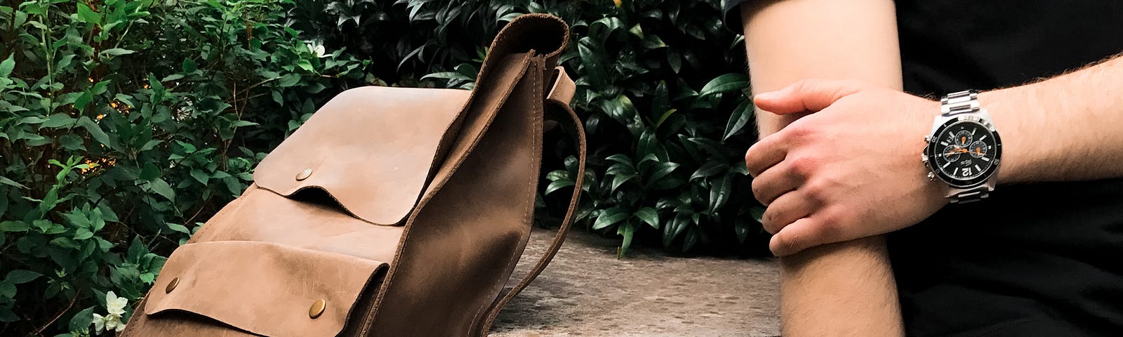 Brown bag on a ledge of a short brick wall next to a person holding their arm with a watch on.