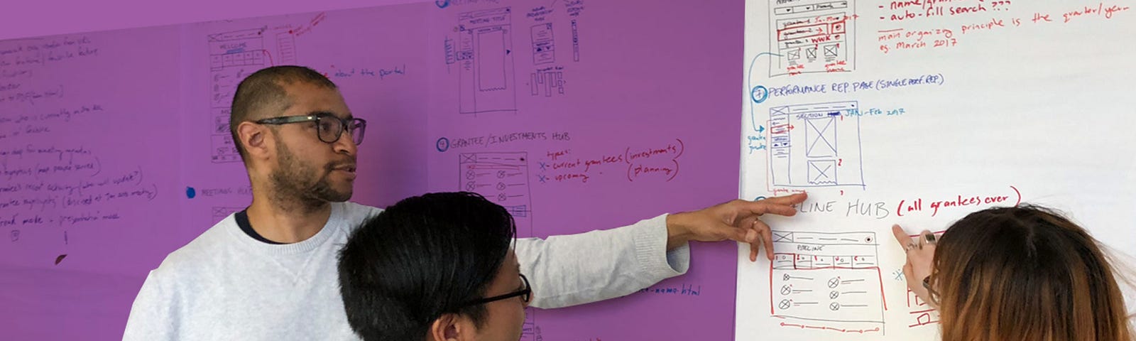 Man pointing at whiteboard full of wireframes with another man and woman looking at what hes pointing at.