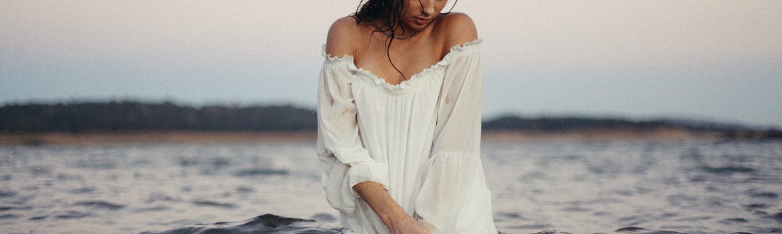 A woman in a white dress swimming knee-deep in ocean at sunset.