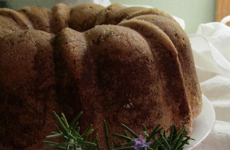 Cake baked in bundt pan on white plate, garnished with sprig of rosemary, in bloom.