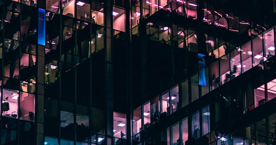 An image of windows of an office building at night with different color (purple, blue, pink) lights illuminating them from the inside.