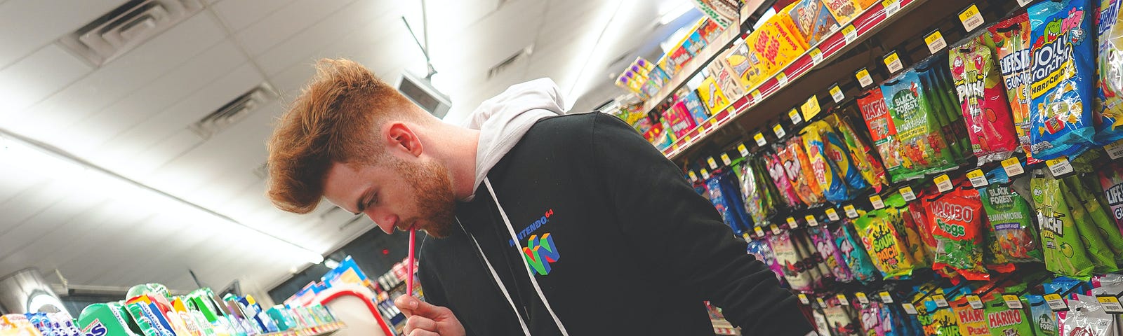 A picture taken on a slant so it feels a bit dizzying. a young man standing in a store between two aisles crammed with packed candies, drinks, etc. He looks like there are way too many choices
