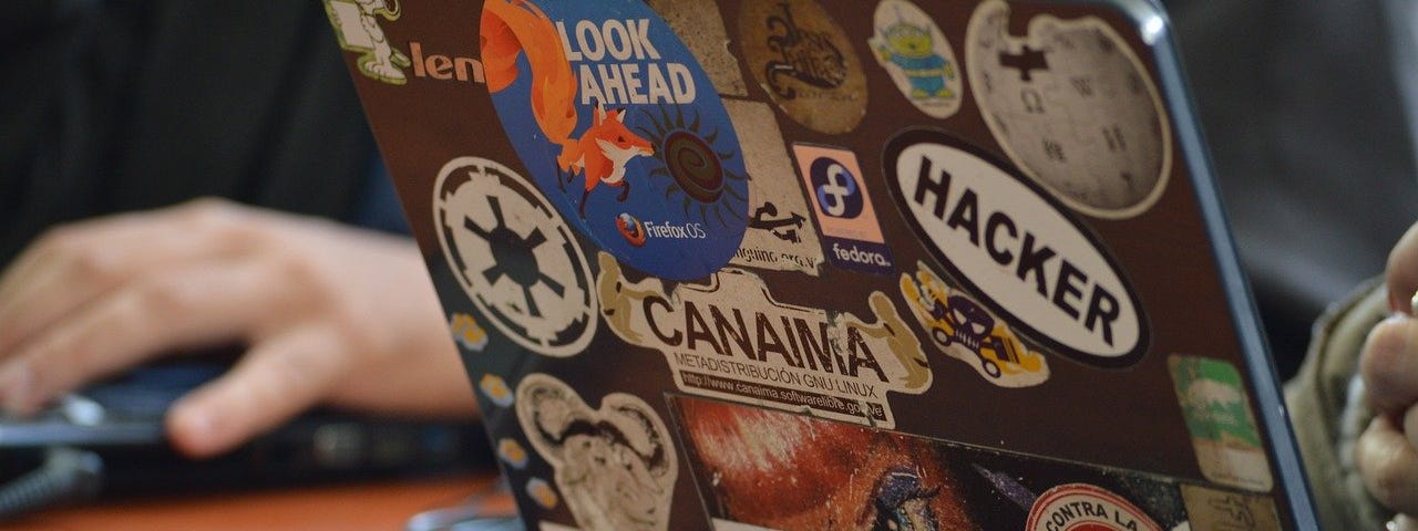 IMAGE: An open laptop with a lot of stickers, one of them reading the word “hacker”