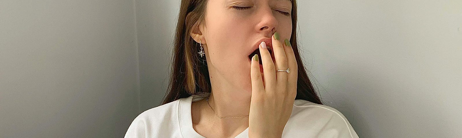 This image shows a young woman sitting in a wicker chair, dressed in a plain white T-shirt with a cute cartoon design. She is yawning and covering her mouth with one hand, while holding a green mug in the other. Her eyes are closed, and she has a relaxed, casual posture. The setting is minimalist with a light gray wall background. She has brown hair, a subtle earring, and a visible tattoo on her arm.