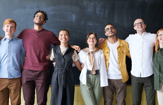 A group of diverse people, men and women, black and white, smiling and embracing in a group picture with a chalkboard behind them.