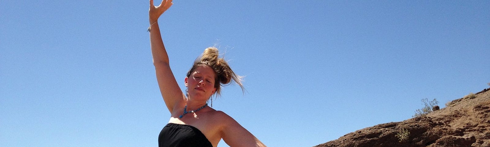 A woman standing on a ledge with blue sky and mountain behind her. Her eyes are closed, the wind blowing her dress and blonde hair. Her hair is in a loose bun. Her left arm is raised to the sky, the right arm lifted slightly from her side. She is wearing a strapless black sundress.