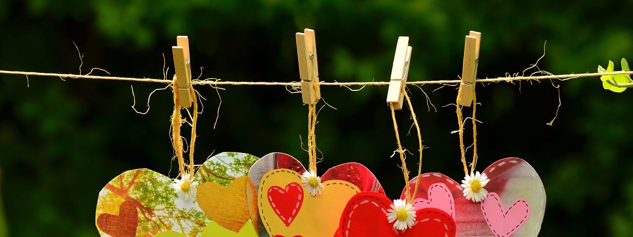 Four clothespins holding four hearts on a string. The first heart on left is in orange and bright green, the second is orange with small red hearts, the third is a red heart with white stitching and the fourth is gray and red with small pink hearts on it. Green grass and trees are in the background.