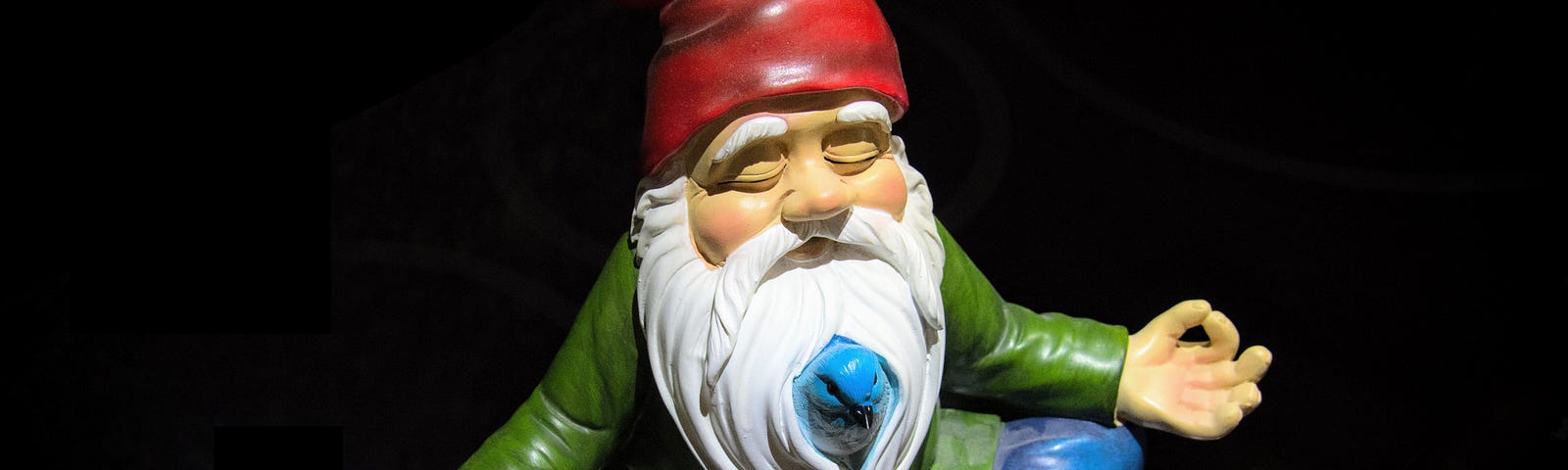 Garden gnome in blue pants, green jacket, red cap, long white beard, sitting cross legged with hands in meditation pose.