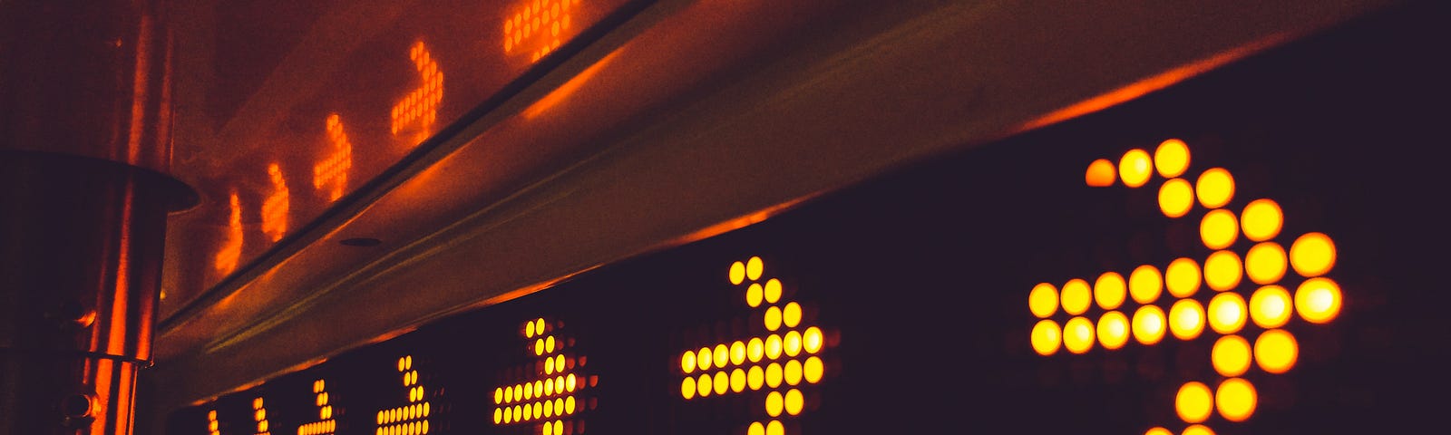 Series of neon yellow lights in the shape of arrows pointing to stage right. There are seven arrows in groups of three. Their reflections can be seen above them in an orange-red color.