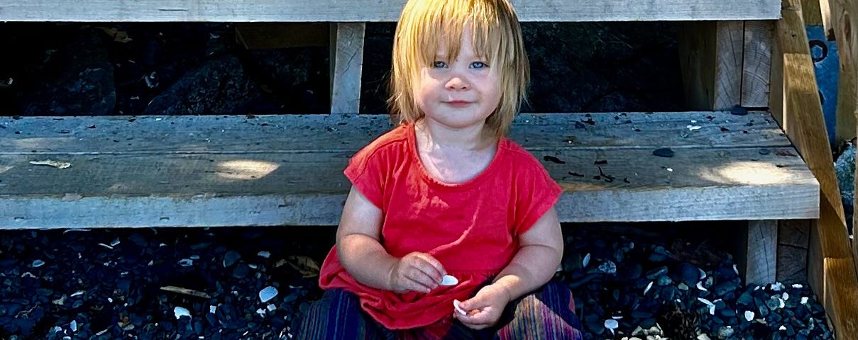 A toddler sits on some steps that lead to a gravel beach. The child is wearing brightly coloured clothes and is holding a shell