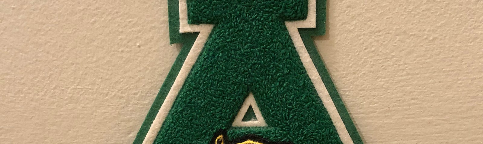 A green letter “A” made of felt, with a patch depicting the lamp of knowledge sewn on it and a gold pin depicting the lamp of knowledge pinned to it