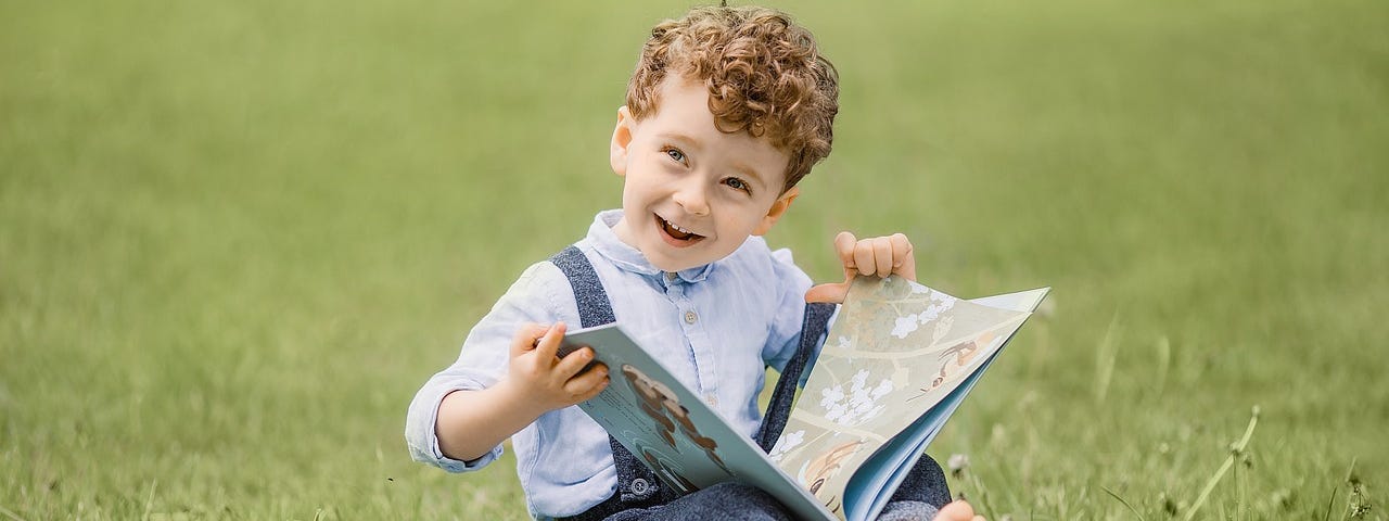 A young boy, sitting on green grass, woth a book in his lap. As he turns to a new page, he looks at someone outside the picture, with pure joy on his face.