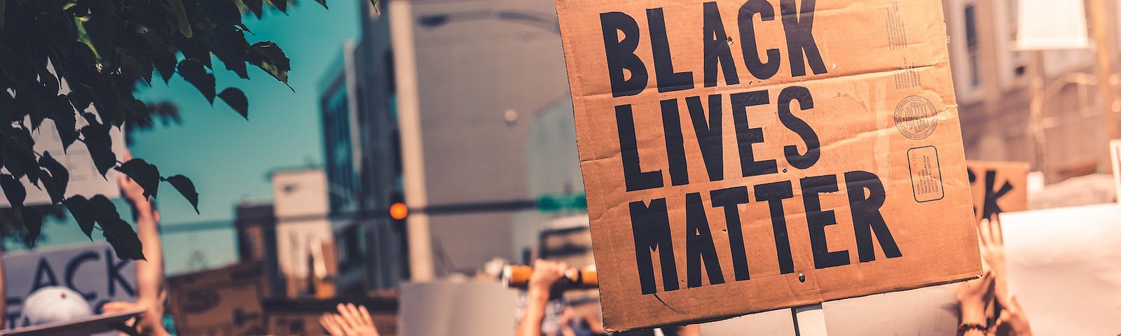 Protestors crowd a street, facing away from the camera. Facing the camera is a cardboard sign saying Black Lives Matter.