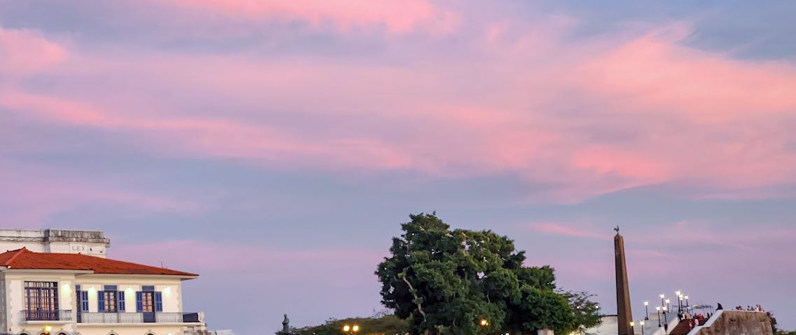 Pink and blue sunset sky over a peninsula with a pillar monument, trees, a lighted walkway, and antique-looking large white building.