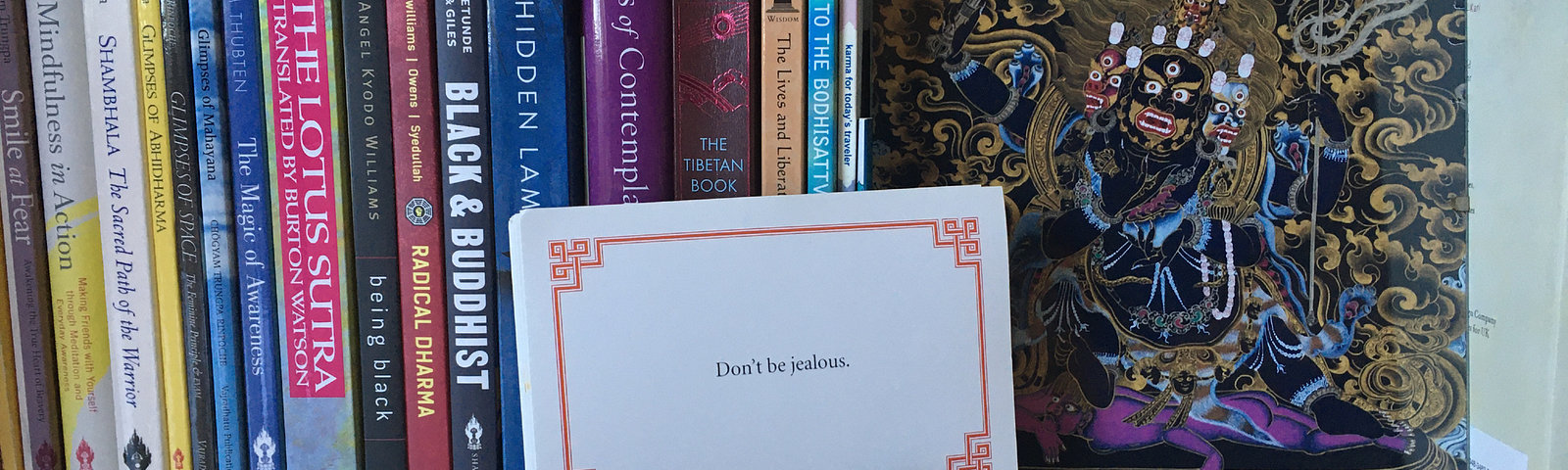 Lojong card that reads, “Don’t be jealous,” sits on a bookshelf in front of some books and an image of a four-armed Mahakala.