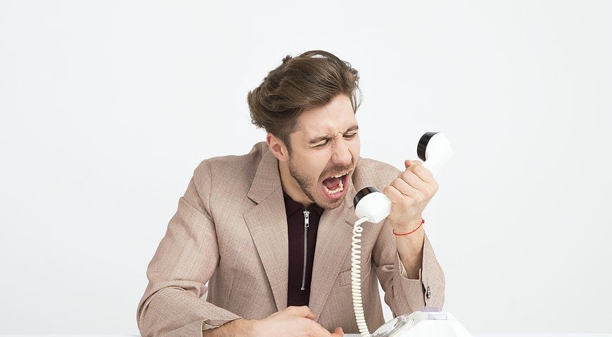A man wearing a business suit yells into a landline phone, with his face contorted by rage