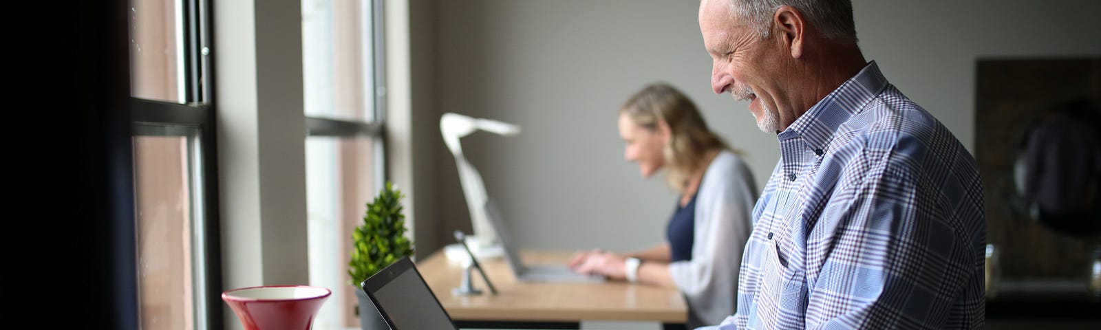 Stock image of two people at standing desks working on laptops. The person in the foreground is an older man. The person in the background is a younger woman.