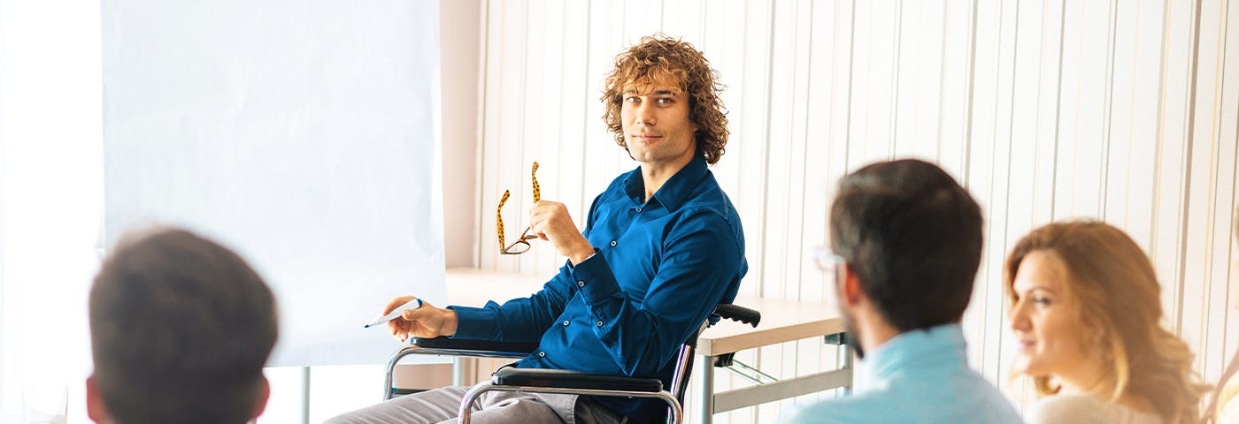 In a professional training session, a curly-haired male instructor sits in a wheelchair in front of a flipchart.