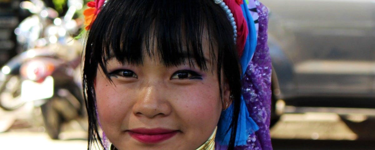 The head and shoulders of a young black-haired, Asian woman smiling directly at the camera. Her neck and upper shoulders are bound in many brass rings and she has colourful headwear on her head.