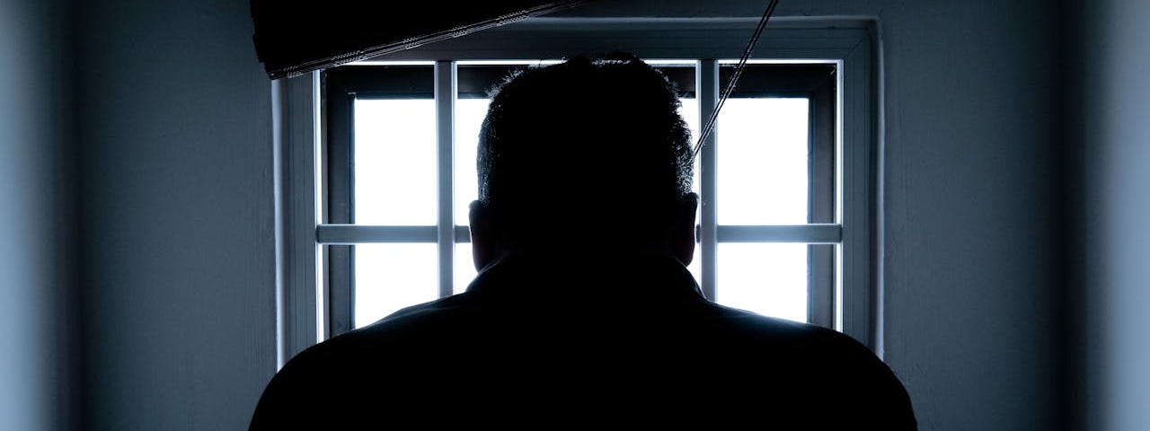 The dark silhouette of a man looks out a small barred window.