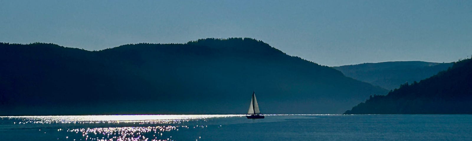 A small sailboat appears across an ocean with hills behind it. The water ripples with sunshine.