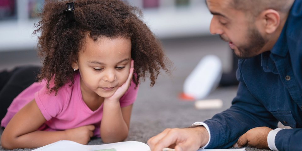 A young girl sits with an adult, reading an open book.