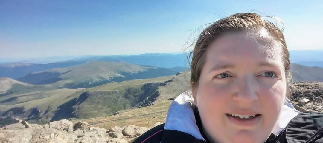 Attractive white woman with pretty eyes and medium brown hair tied back in a ponytail stands atop a mountain with other mountains and clear blue sky behind her.