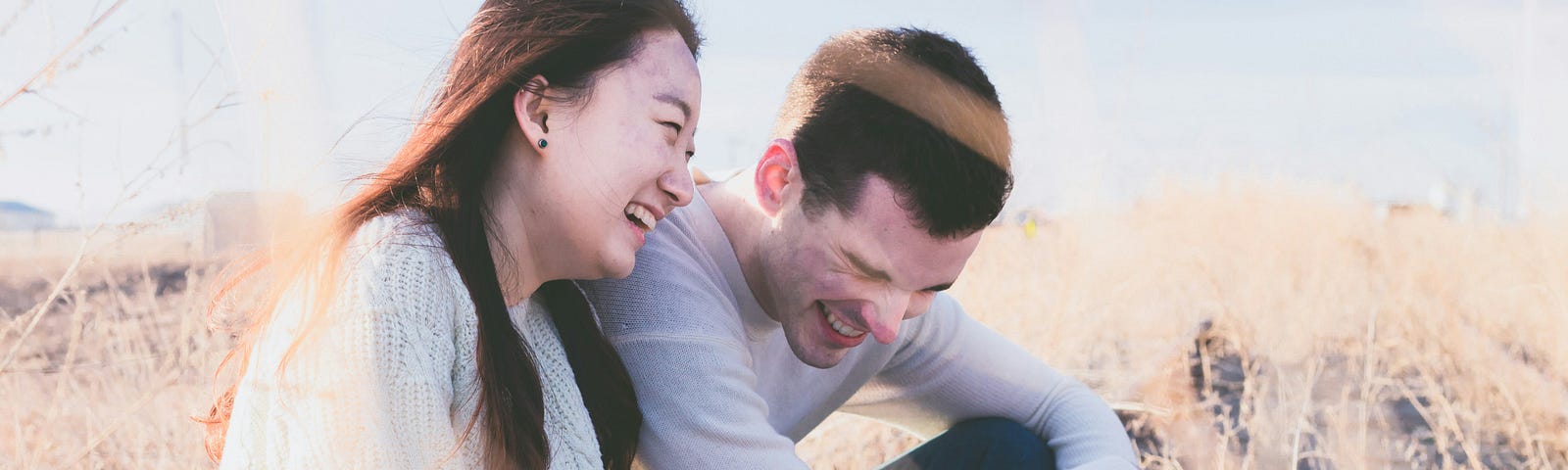A man and woman sit laughing in a field under clear skies.