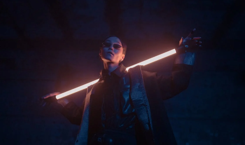 Person in black suit and dark sunglasses holding a neon sword.