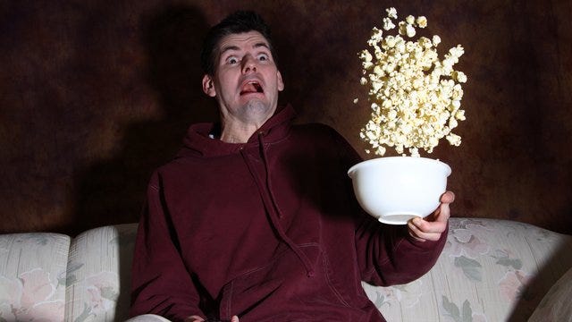 Image of a white man seated on a sofa, an exaggerated expression of terror on his face. He is holding a bowl of popcorn and the image captures it in motion, having been tossed into the air.