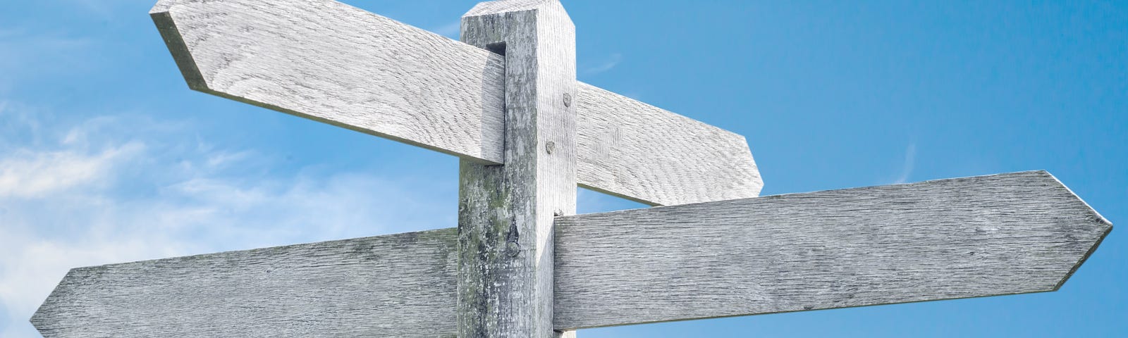 A light grey wooden signpost with four directional boards, without any words or symbols on it. There is a blue sky with wispy clouds in the background.