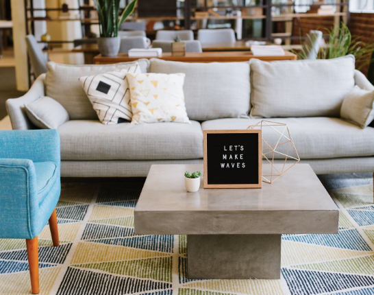 A coworking space featuring a couch, an armchair, and a concrete coffee table with a sign reading, “let’s make waves” atop it.