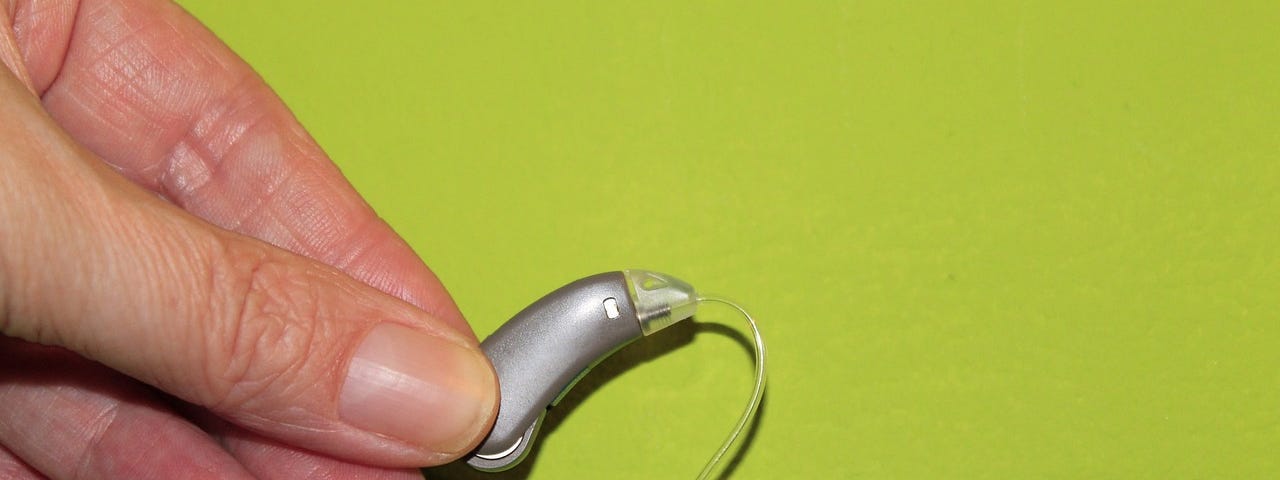 IMAGE: On a lime green background, a hand holding a hearing aid device