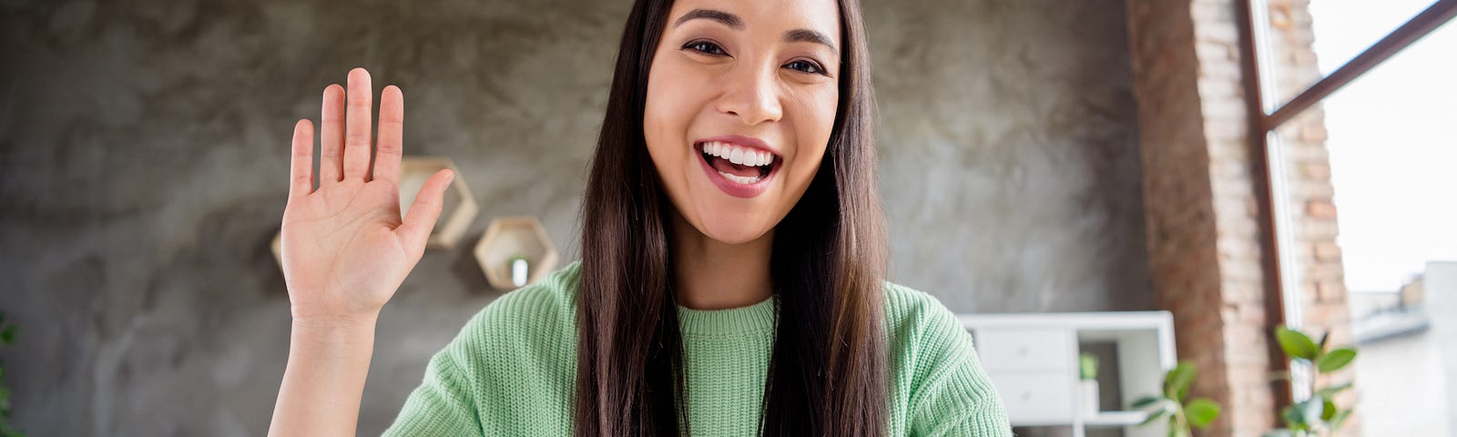 Close-up of a cheerful Asian female working from home and waving at her laptop’s webcam