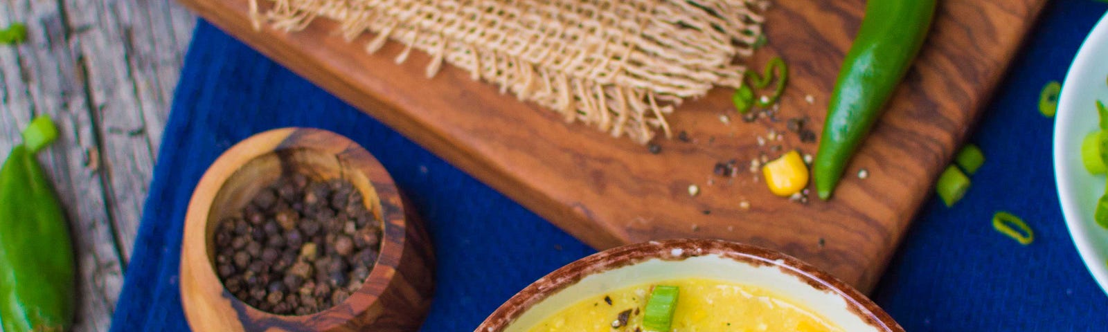 yellow corn chowder with green onions and fresh cracked black pepper, next to a blue tablecloth and peppercorn bowl